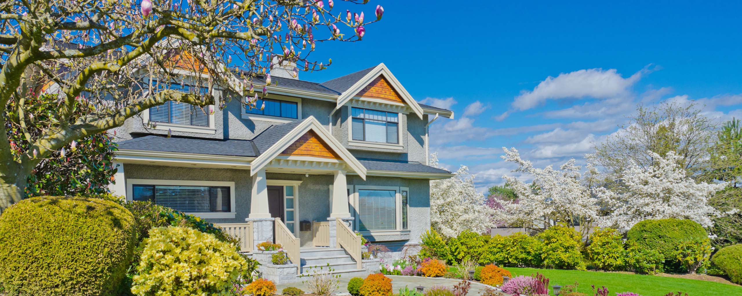 A two-story house with a blue sky backdrop is surrounded by blooming trees and vibrant spring flowers - https://murphyinsgrp.com/